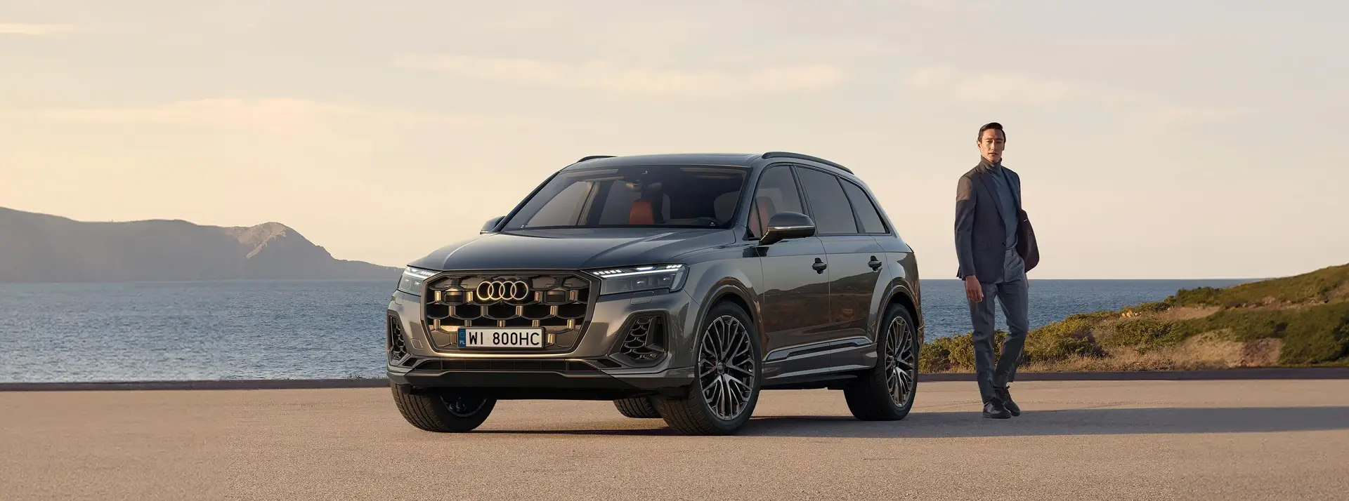 parking, w oddali góry, mężczyzna stoi obok ciemnoszarego SUV-a Audi SQ7