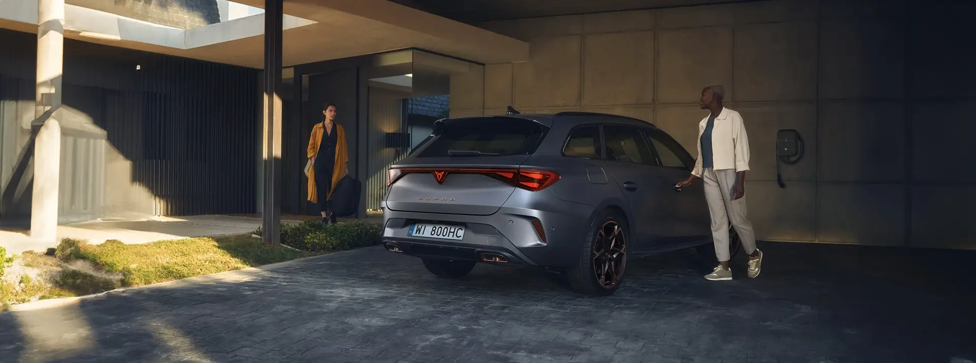 szare auto CUPRA Leon Sportstourer zaparkowane przed nowoczesnym domem, obok stoją dwie kobiety